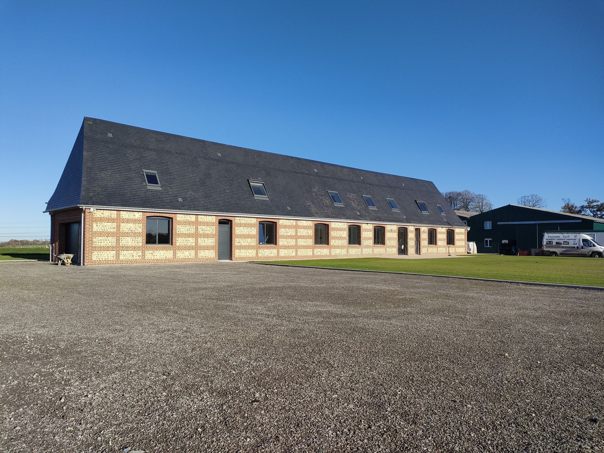 Long bâtiment en construction, aux murs lambrissés et au toit sombre ; terrain gravillonné et ciel bleu azur.