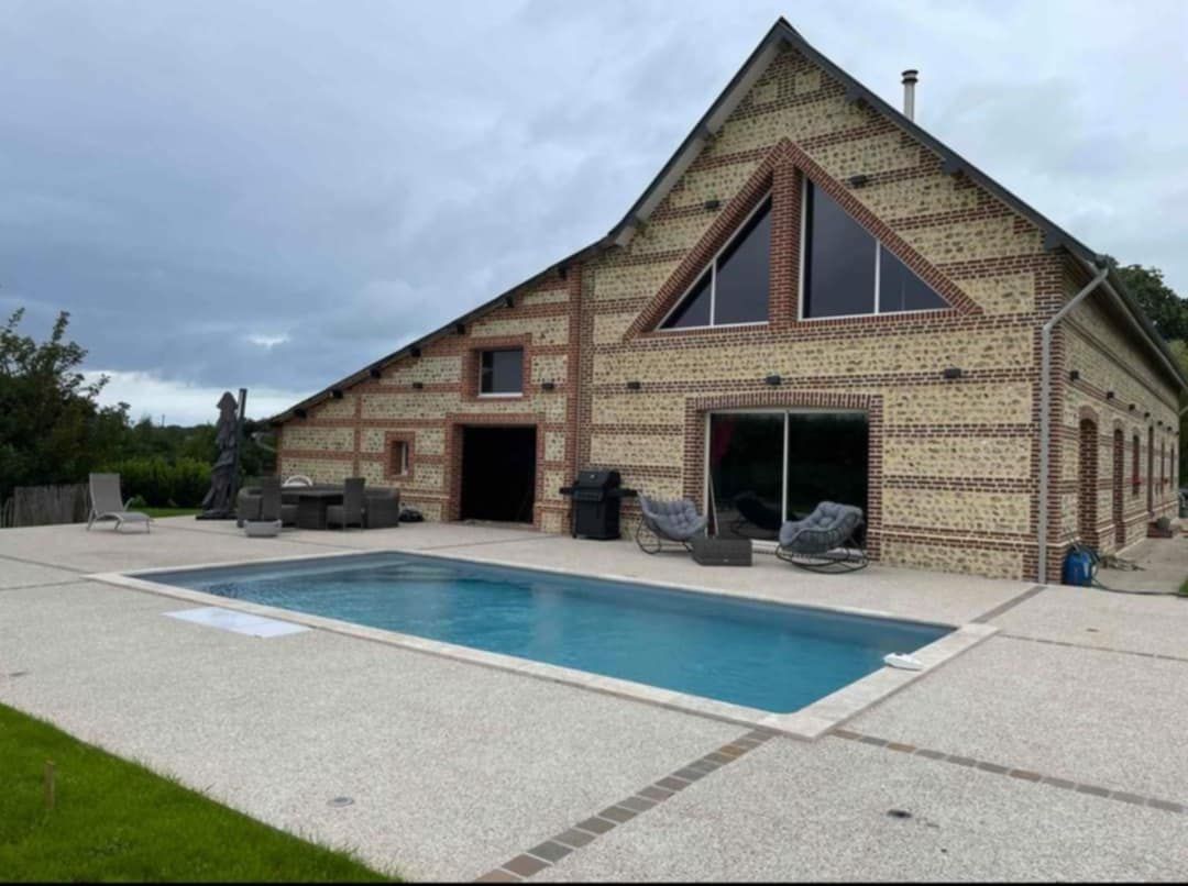Maison moderne avec piscine, façade en briques et en bois, sous un ciel nuageux.