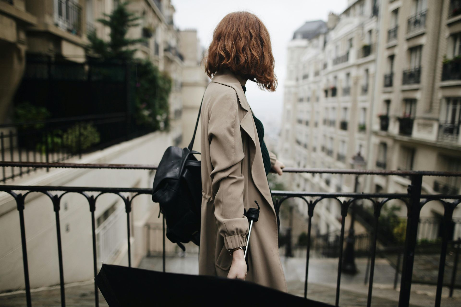 Une femme vêtue d'un manteau beige, portant un sac à dos noir et tenant un parapluie, se tient sur un pont surplombant un paysage urbain européen.