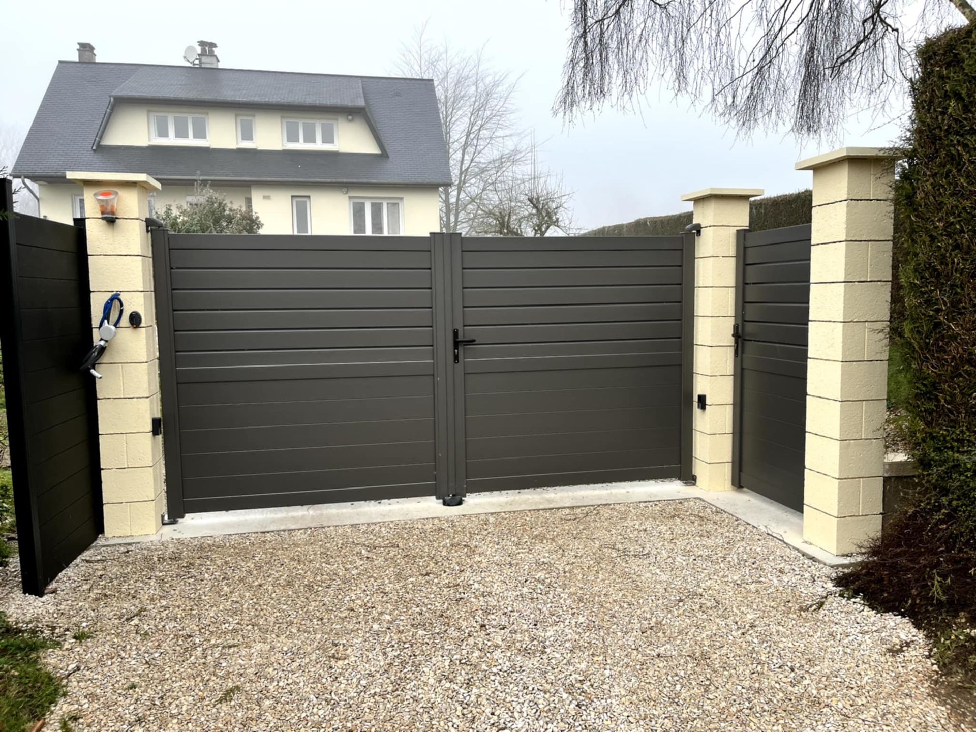 Un portail en métal gris se dresse devant une maison. Il est encadré par deux piliers beiges et une allée est recouverte de gravier.