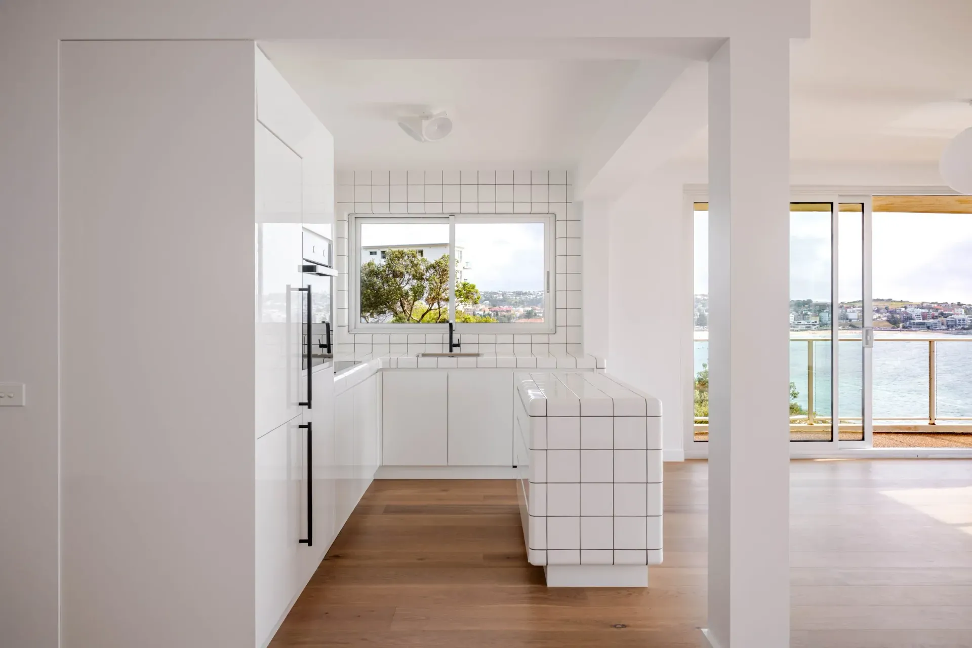 Bright modern kitchen with white cabinets, wood floors, and ocean-view windows opening to a balcony.