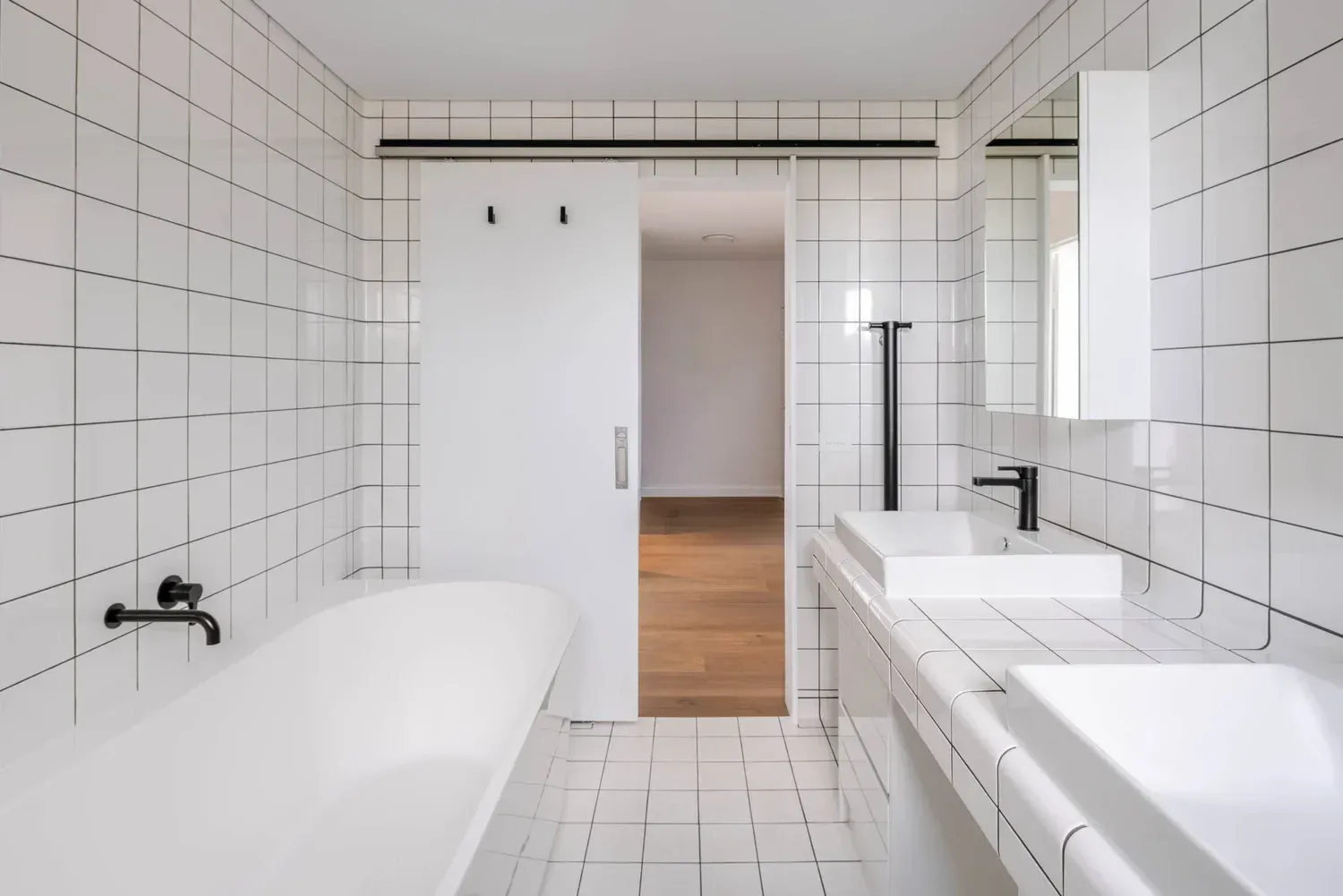 Bright modern bathroom with white tile, freestanding tub, sink, and open doorway to wood floor room