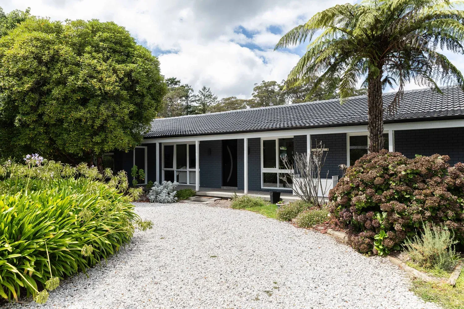 Single-story house with white gravel driveway, lush garden, and palm trees under a cloudy sky