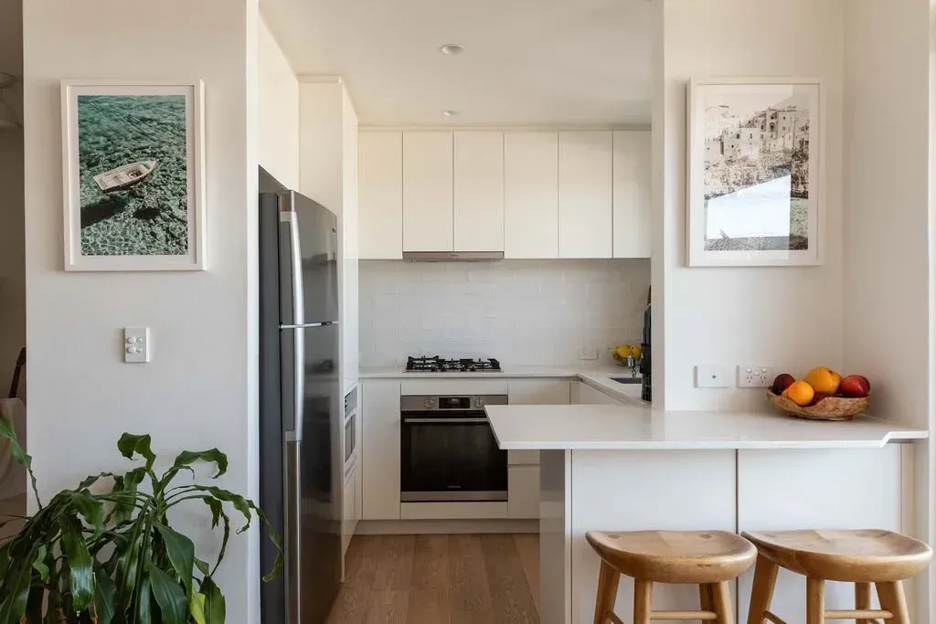 Bright kitchen with white cabinets, stainless stove, breakfast bar, stools, and a leafy plant by the wall.