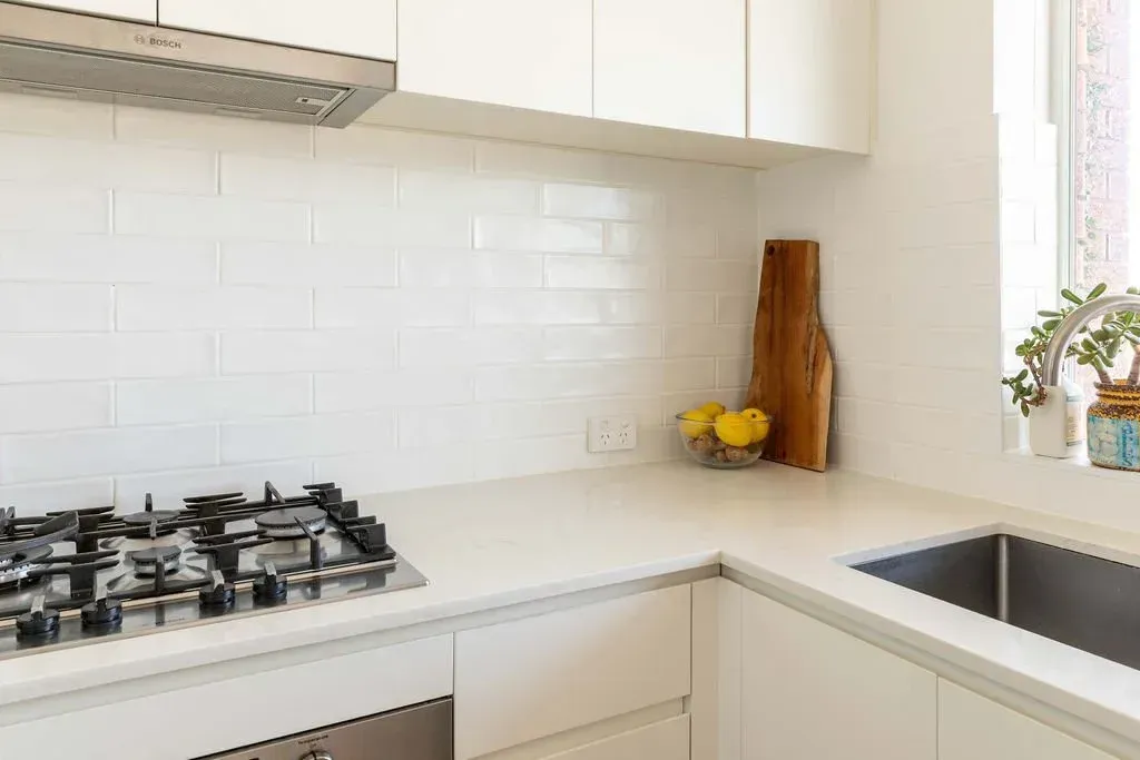 Bright white modern kitchen with gas stove, cabinets, sink, and wooden cutting board.