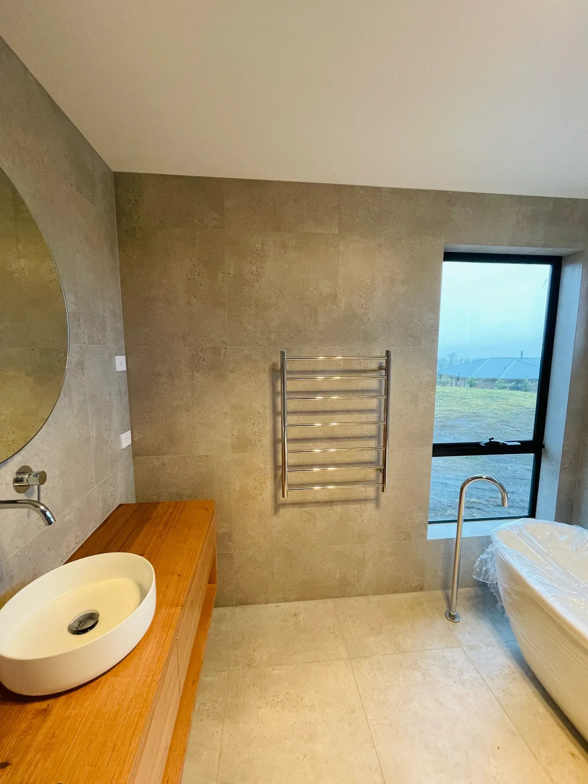 Modern bathroom with a wooden vanity, vessel sink, wall-mounted heated towel rack, and a freestanding tub by a window. — Hobart Tiling Solutions In Clarendon Vale, TAS