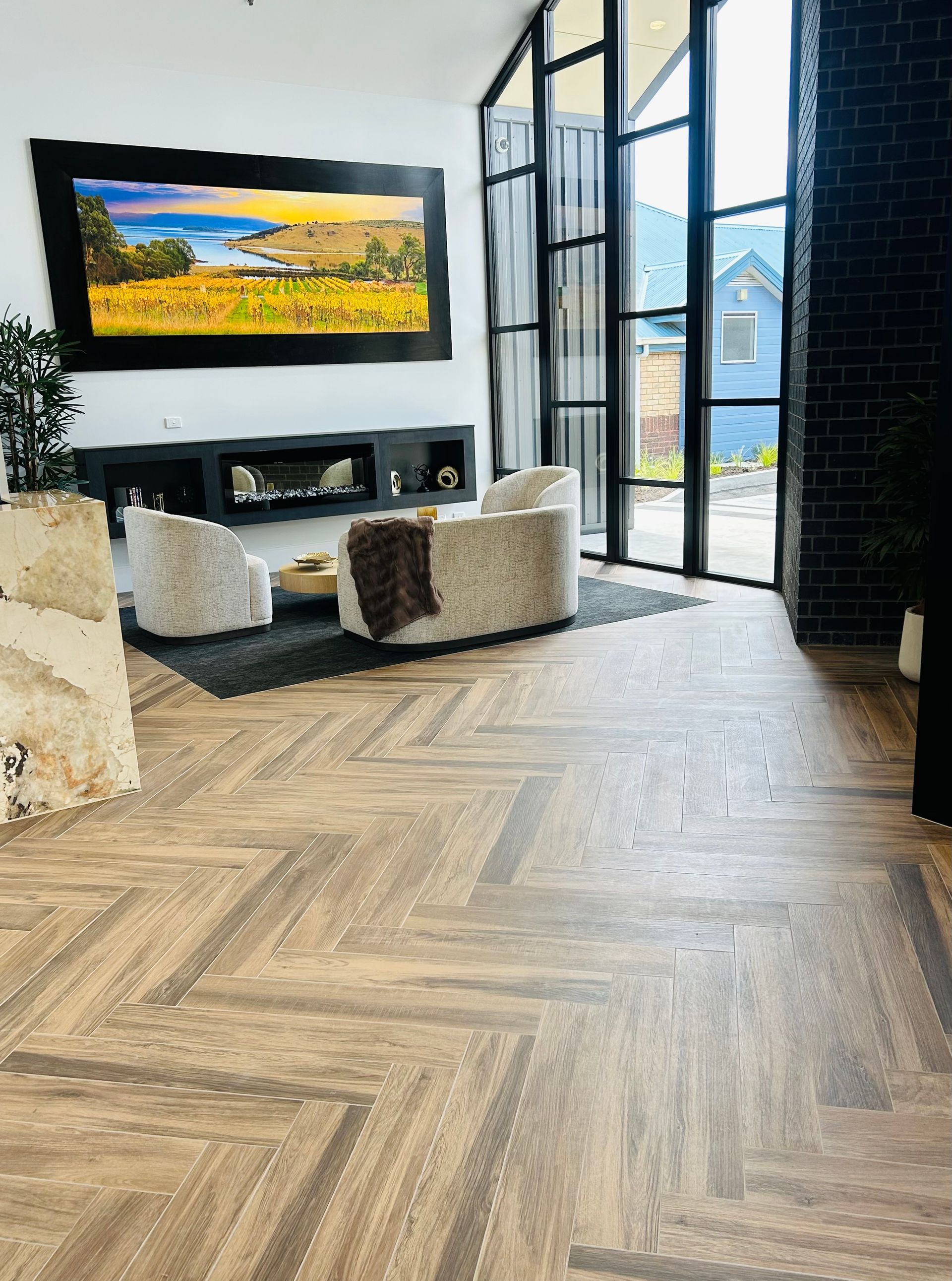 A modern living room with herringbone wood floors, two beige armchairs, a fireplace, and a large landscape painting.  — Hobart Tiling Solutions In Clarendon Vale, TAS