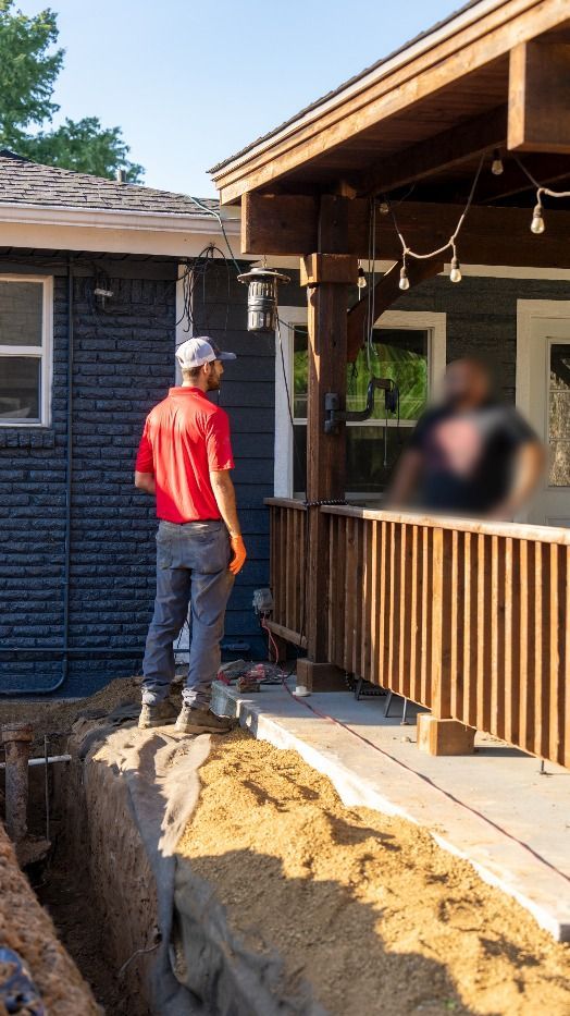 Man in red shirt stands near trench by a porch, looking toward it. Exterior construction scene.