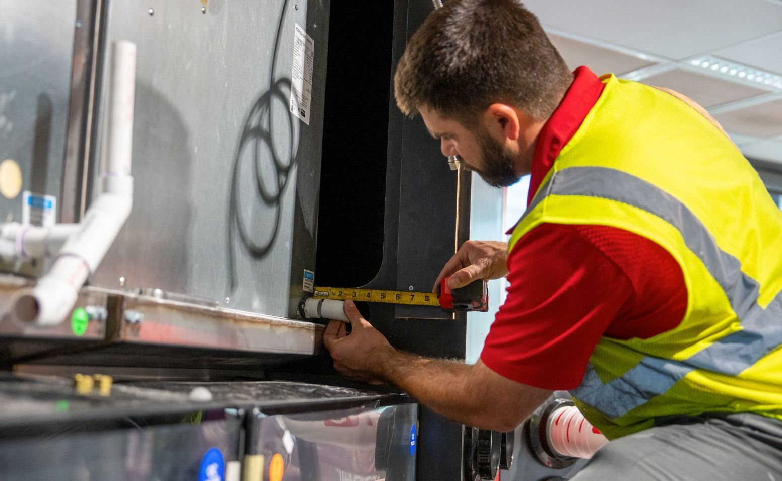 A worker in a yellow vest measures a pipe with a tape measure on machinery.