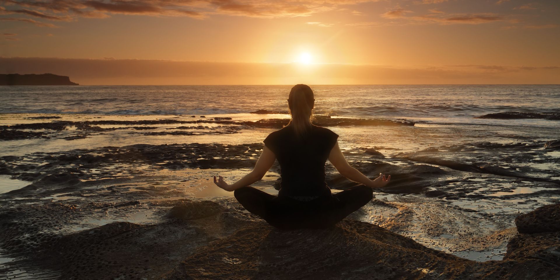 A woman is sitting in a lotus position on the beach at sunset.