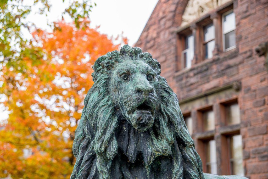 Bronze lion statue with open mouth, autumn trees, and brick building in the background.