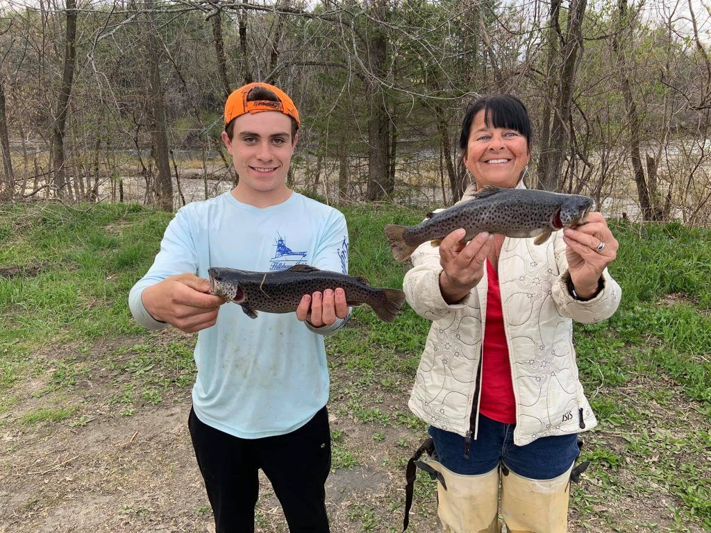 Two people holding up fish. A young man and older woman smile outside. Trees in the background.