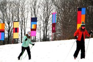Two people cross-country ski in the snow, passing colorful vertical art installations.