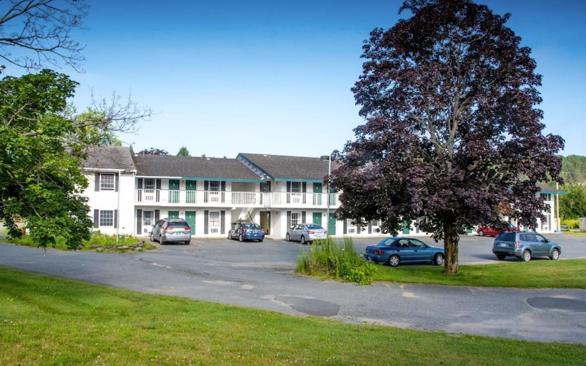 Motel with parking lot and cars, green lawn, and a large tree.