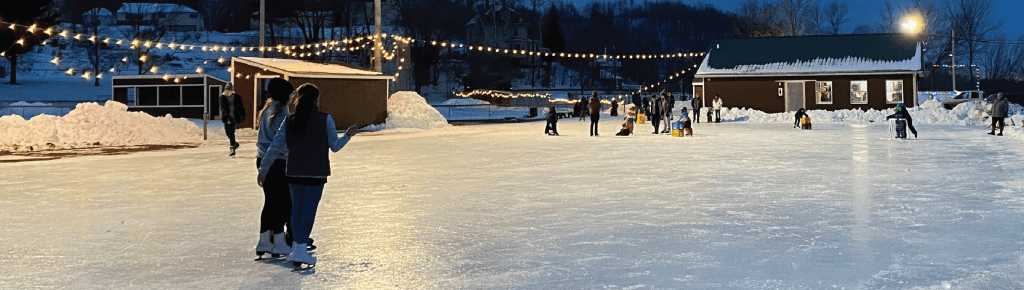 People ice skating at an outdoor rink at dusk, with string lights, snow, and buildings.