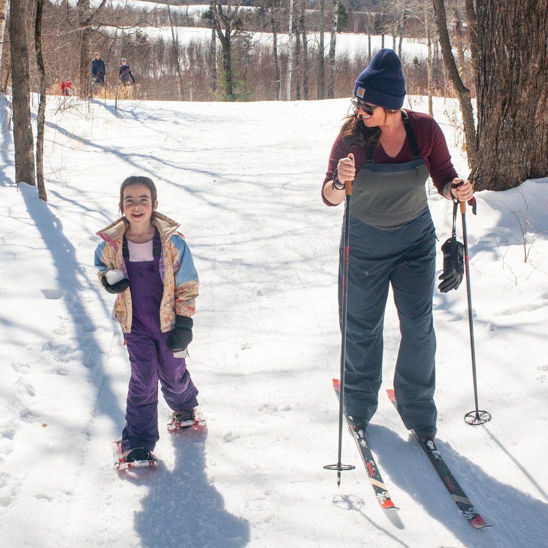 A woman skis, and a child in snowshoes stands in the snow on a sunny day.