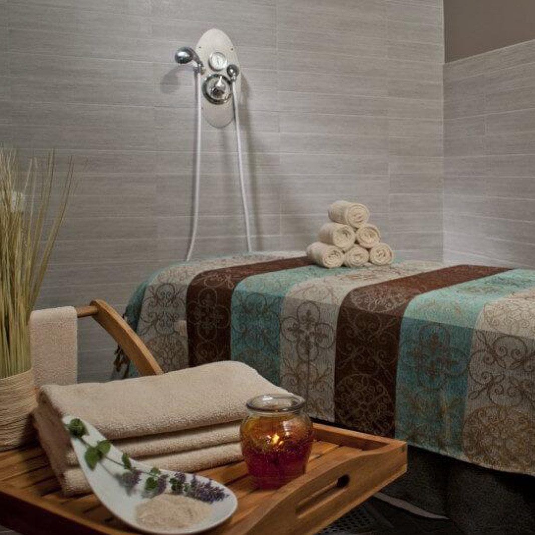 Spa setting: massage table with towels, candles, shower fixture. Serene, muted colors.