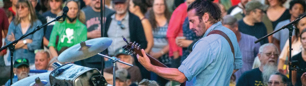 A man playing guitar onstage at an outdoor concert, facing a large crowd.