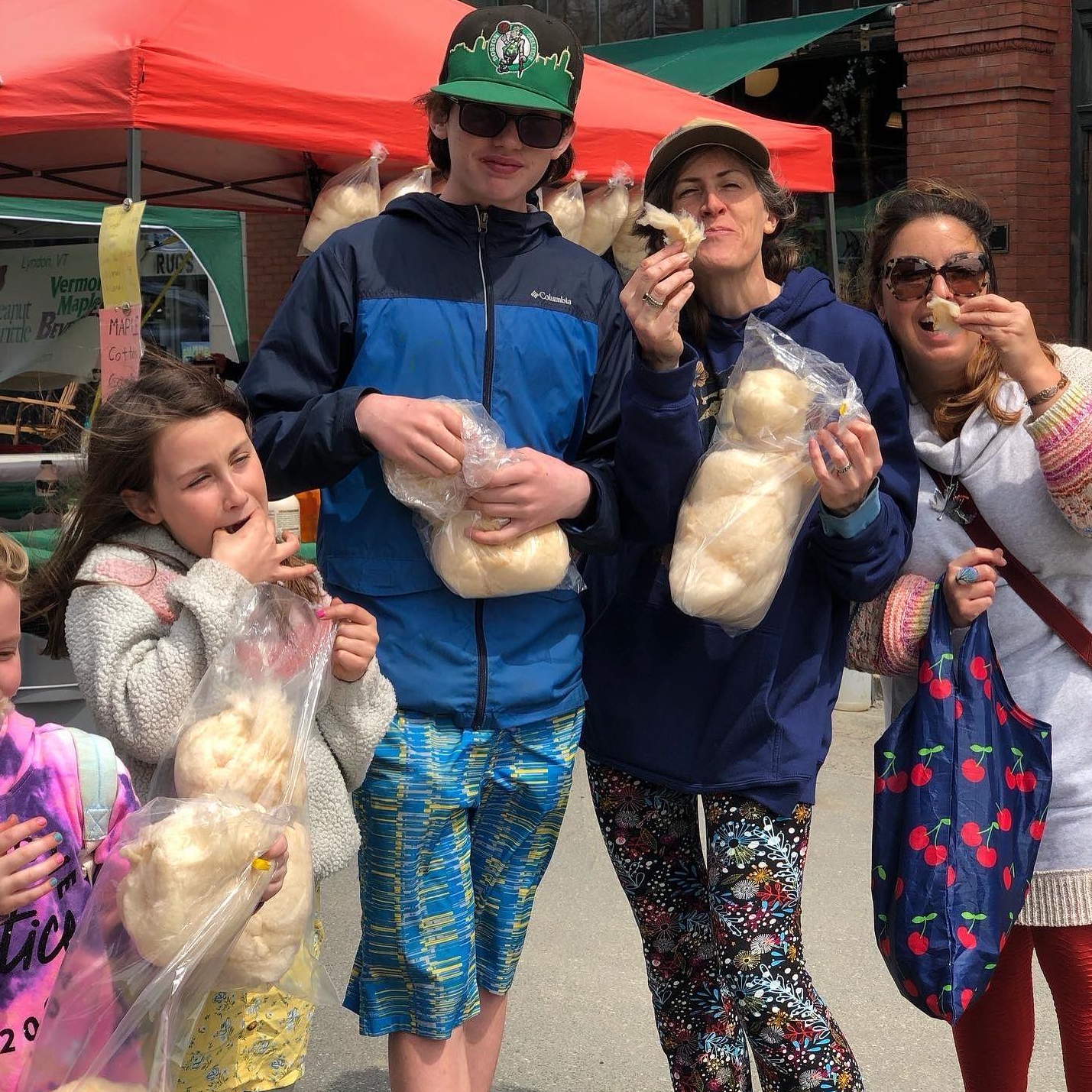 Family eating cotton candy at an outdoor market. People smiling and wearing colorful clothes.