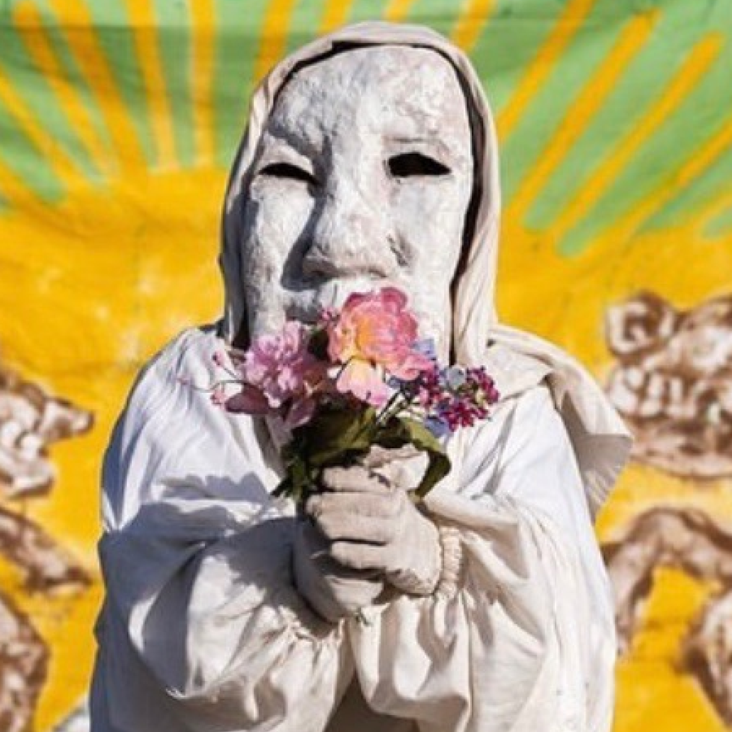 Person in white mask holds flowers in front of yellow painted wall.