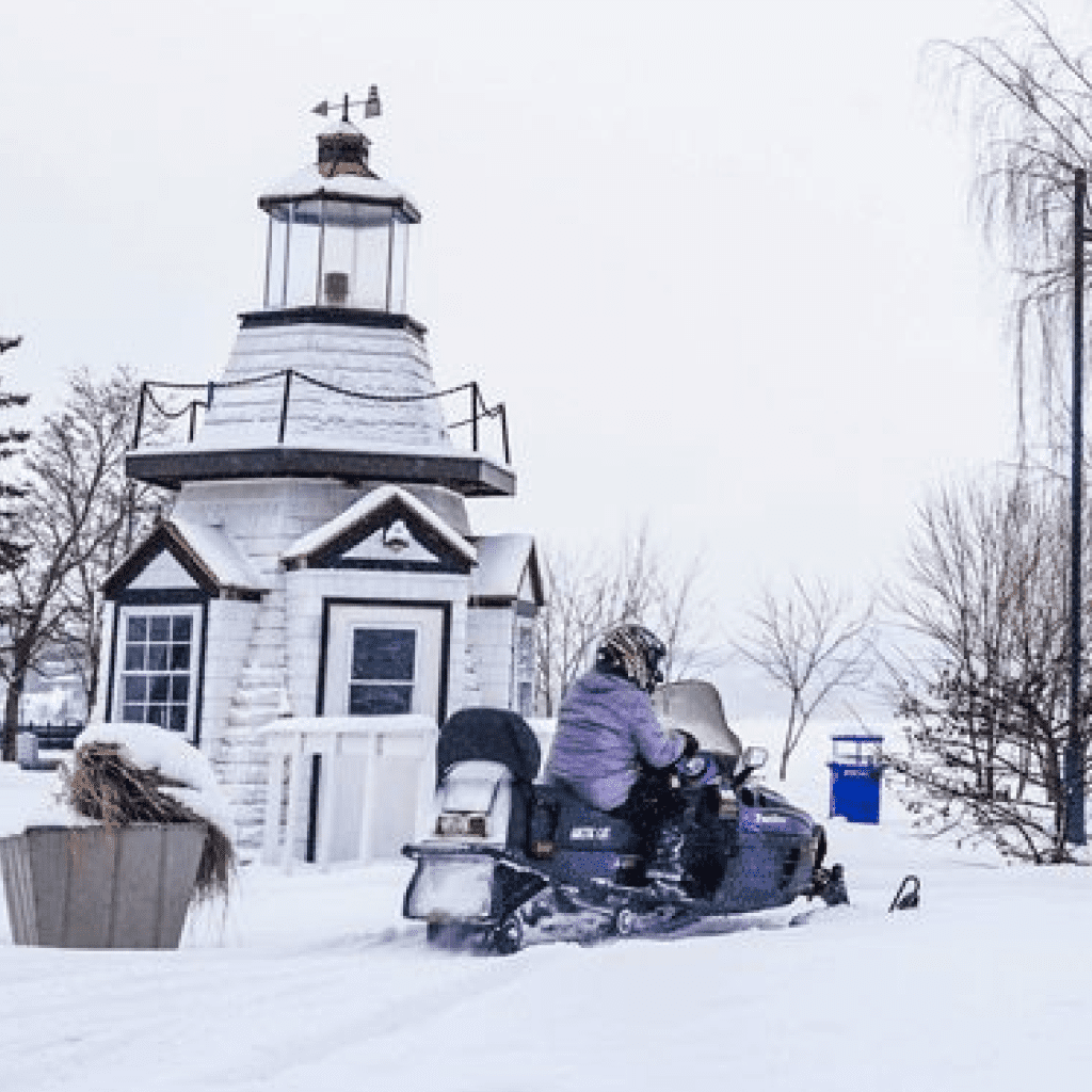 Person on snowmobile in front of a white lighthouse in a snowy landscape.