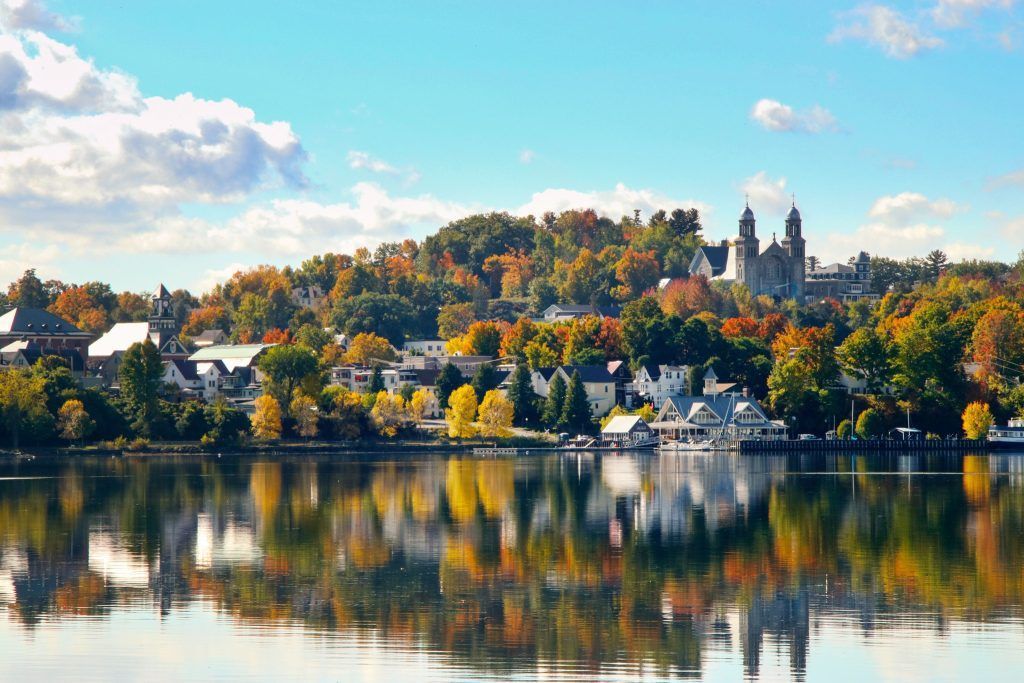 Autumn trees line a waterfront town, reflecting in the calm water. Sunny day with blue sky and clouds.