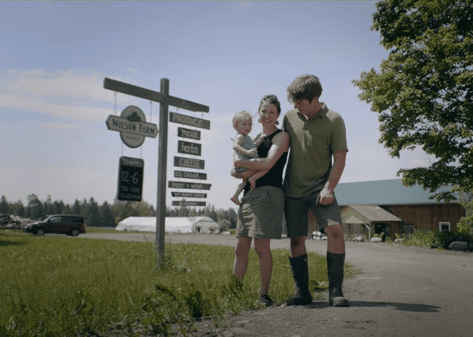 Family of three stands by farm sign on sunny day.