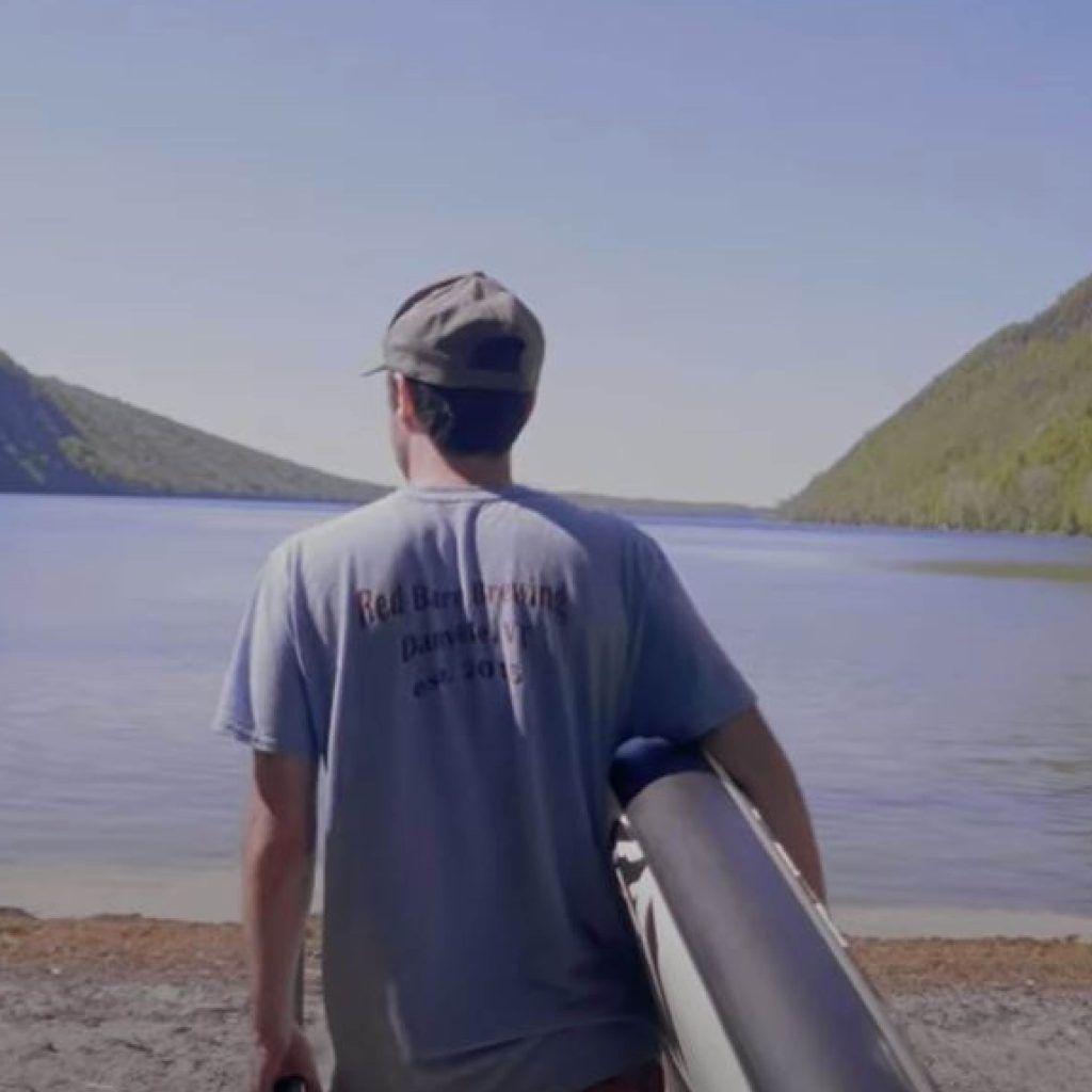Man with a kayak standing at the shore of a lake, mountains in background.