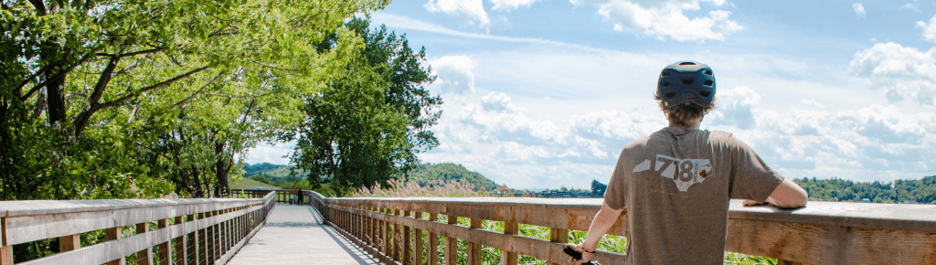 A person in a cap looking out at a lake from a wooden boardwalk. Green trees and blue sky in the background.