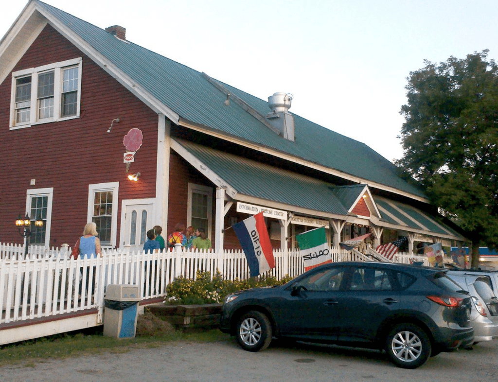 Red building with green roof, white picket fence, car parked out front, flags, and people.
