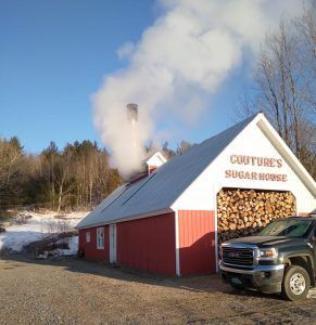 Red sugar house with smoke billowing, a truck parked outside, and a wood pile.