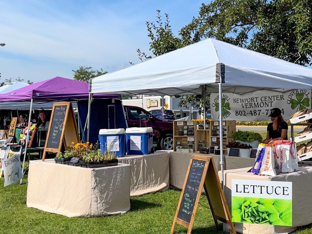Farmers market with white and purple tents on a sunny day; vendors selling vegetables.