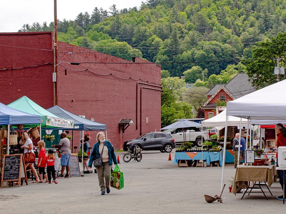 Outdoor farmers market with vendors under tents, shoppers, and a red brick building.