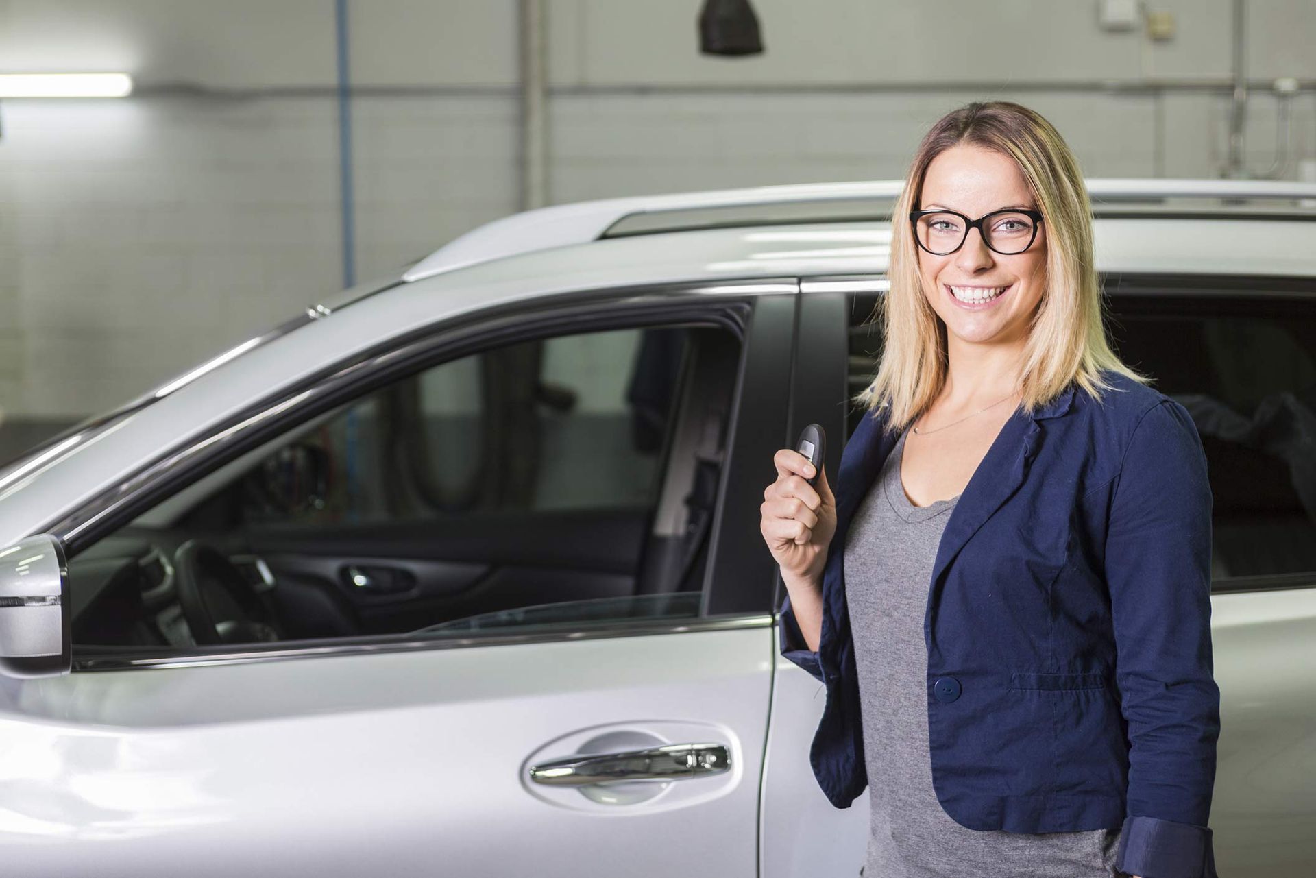 A blonde woman with glasses stands, smiling, next to a silver car while holding its keys. A blonde woman with glasses stands, smiling, next to a silver car while holding its keys.