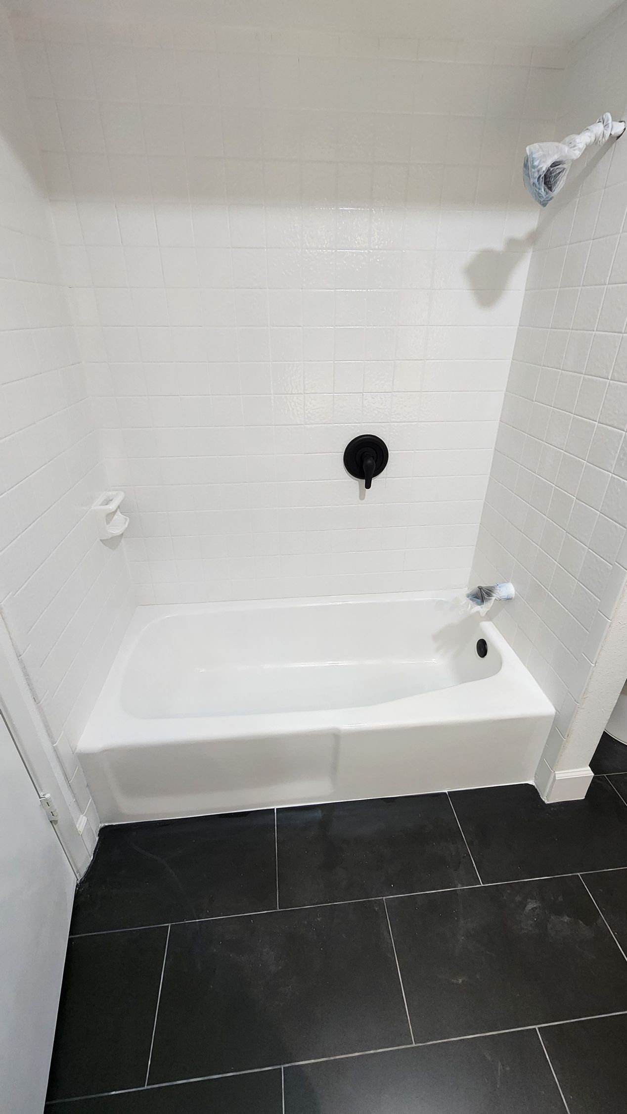 Small white tiled shower with built-in tub, black floor tiles, and grab bars near the faucet.