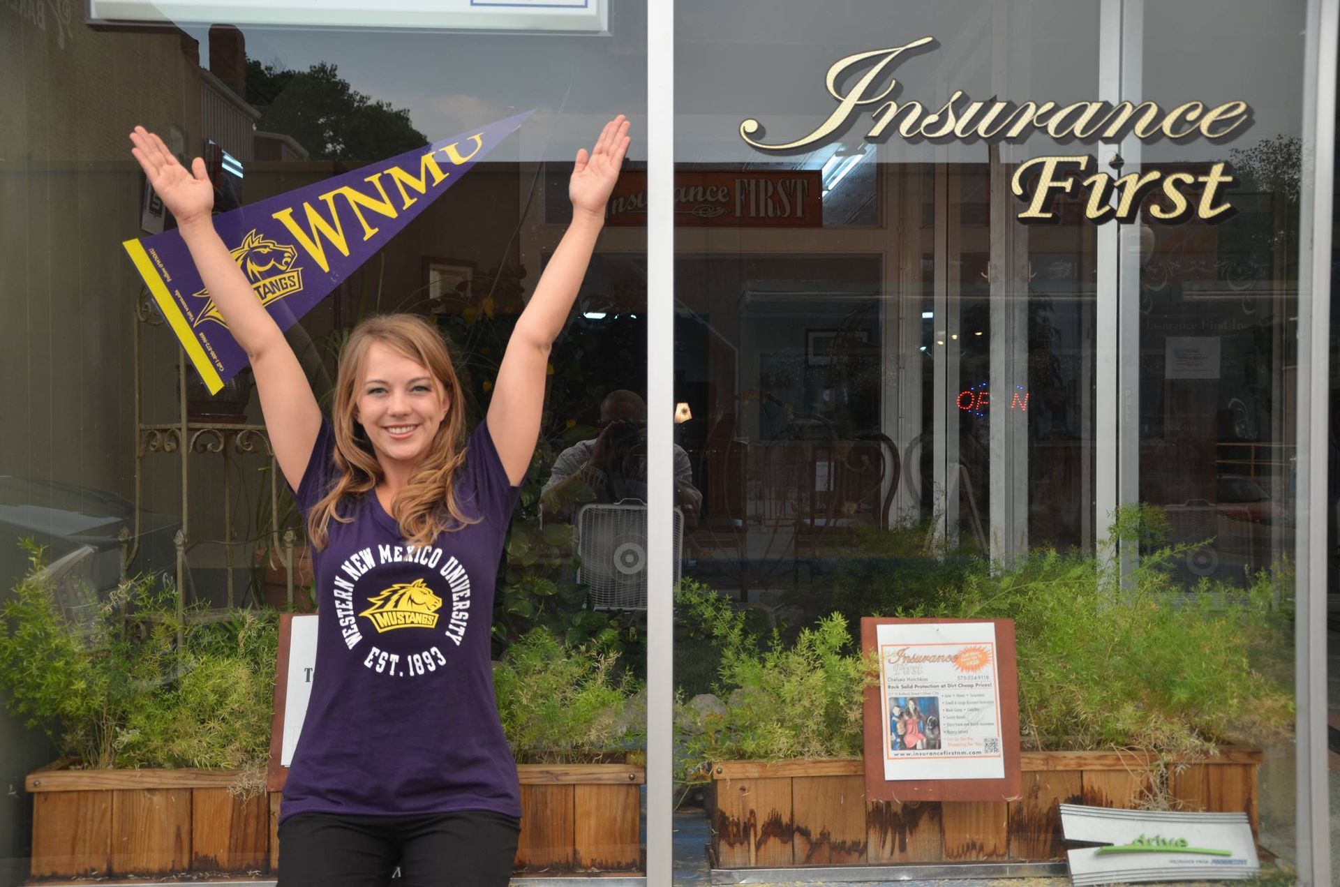 Chelsea in a purple shirt is standing in front of a window that says Insurance First