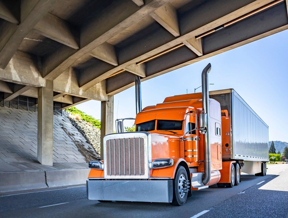 An orange semi truck is driving under a bridge on a highway.