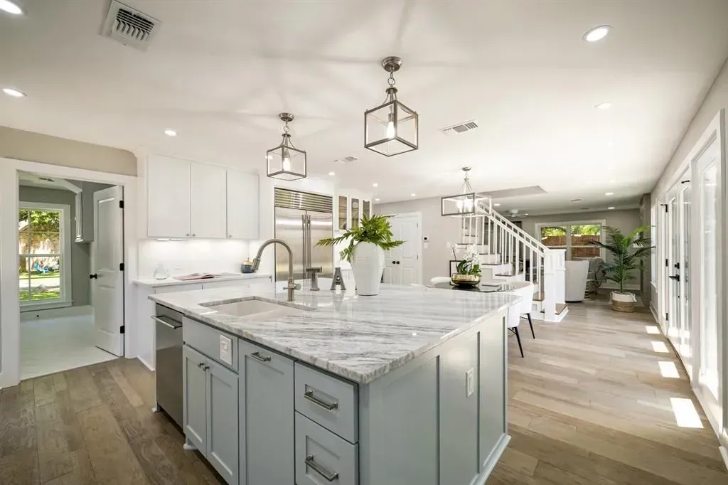 Bright, open kitchen with a large marble island, white cabinets, wood floors, and staircase in the background