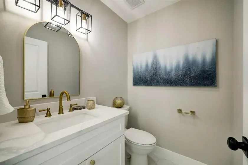 Bright bathroom with gold fixtures, arched mirror, wall art, and toilet beside a white vanity