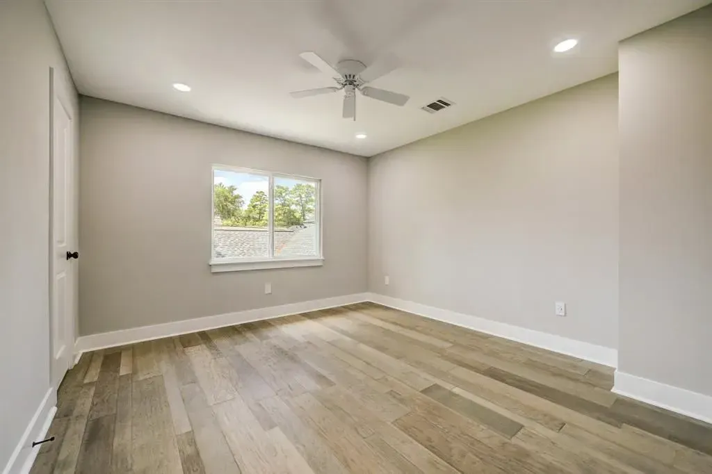 Empty beige bedroom with wood floors, ceiling fan, and a window letting in natural light