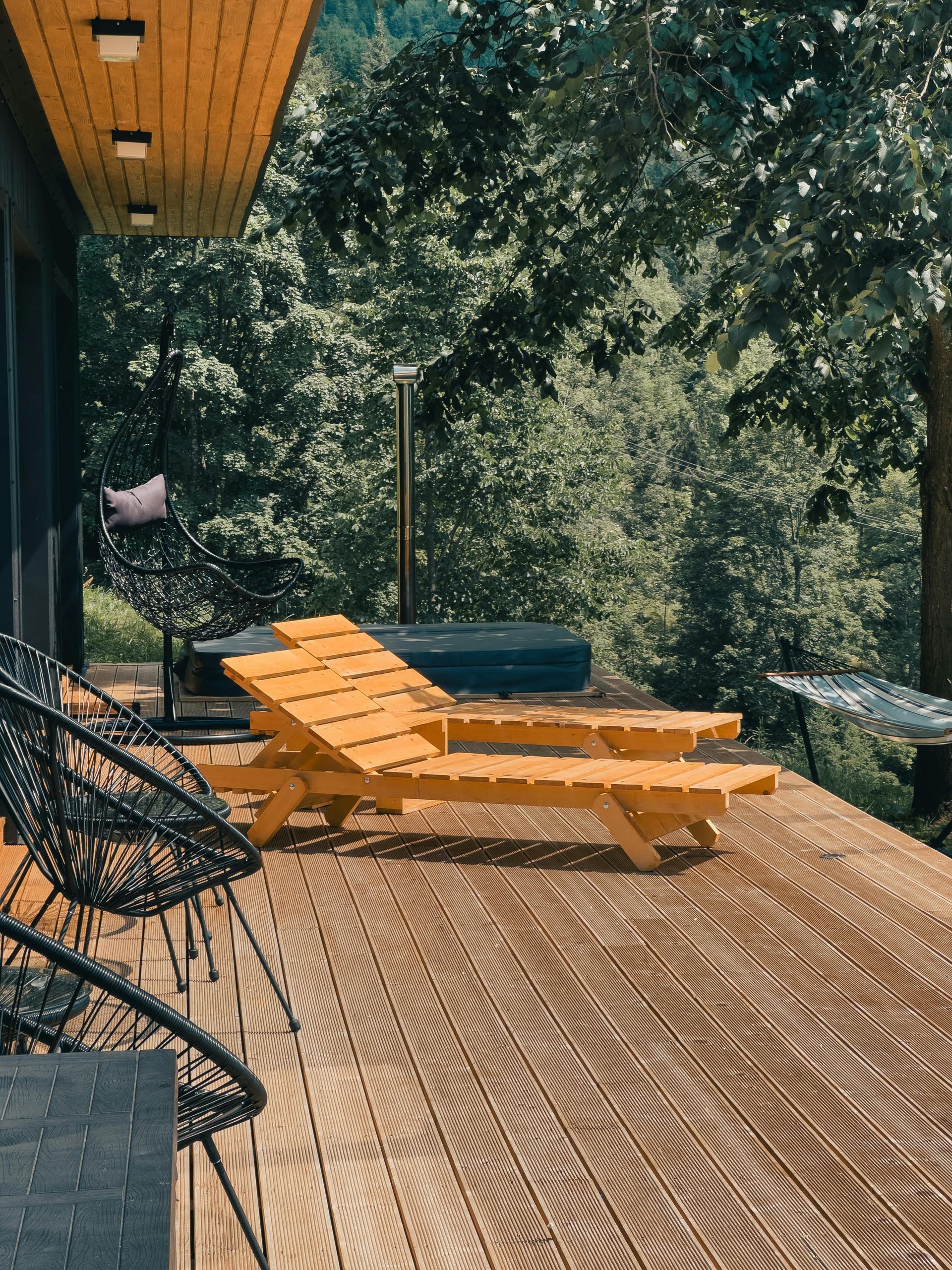 Wooden lounge chair on a sunlit deck surrounded by trees, with wicker chairs and a wooden ceiling overhead