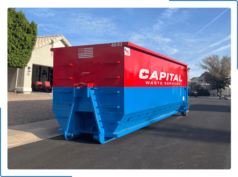 A large red and blue Capitol Waste Services dumpster parked on a residential street under a blue sky.