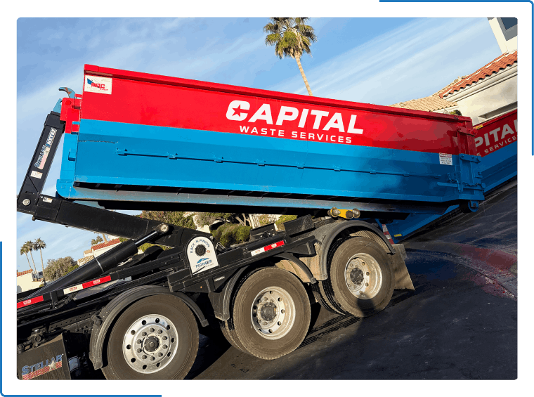 A red and blue Capital Waste Services dump truck trailer being lowered onto the ground outdoors.
