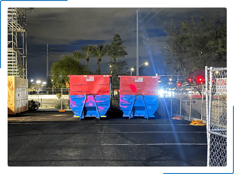 Two red and blue dumpsters sit side-by-side in a parking lot at night with palm trees and a chain-link fence nearby.