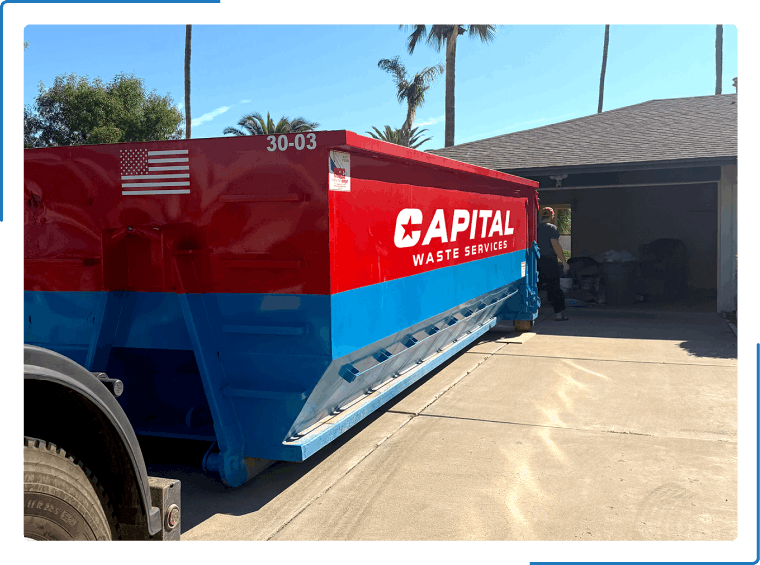 A red and blue Capital Waste Services dumpster parked in a residential driveway next to an open garage.