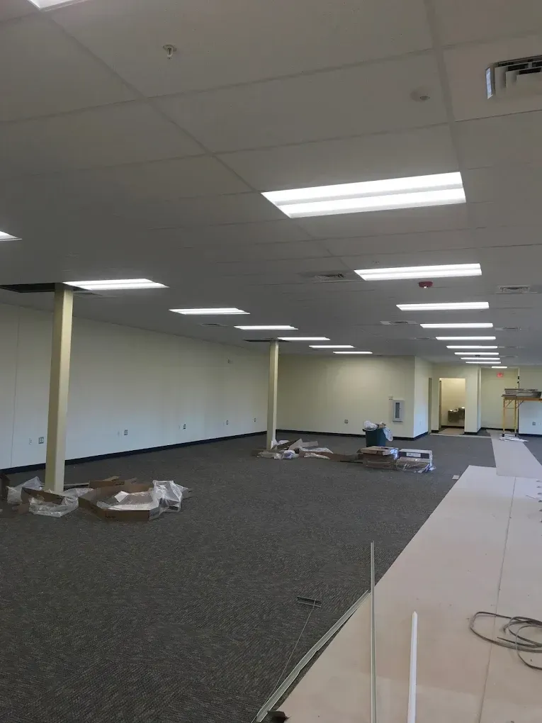 Empty office space with white walls, carpet, and ceiling lights. Construction materials are scattered on the floor.