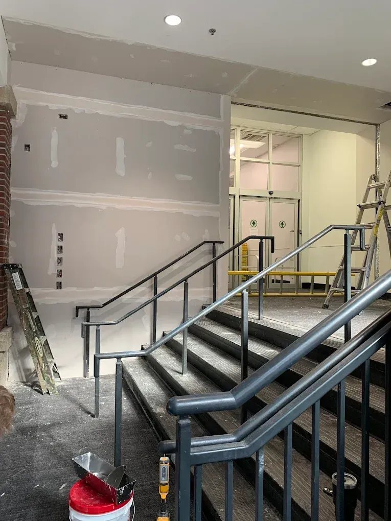 Construction in progress at a stairwell. Drywall, handrails, and a ladder are visible.