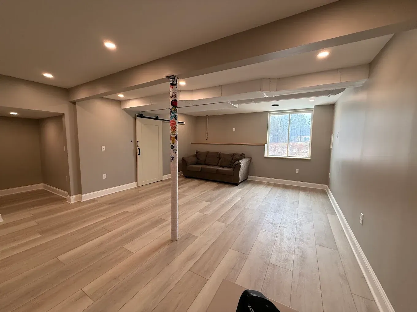 Spacious basement with light wood flooring, grey walls, a brown sofa, and a support pole.