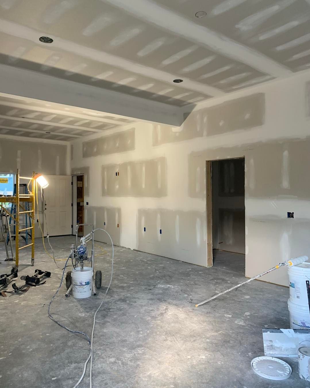 Interior construction site with drywall on walls and ceiling. Tools, buckets, and ladder present.