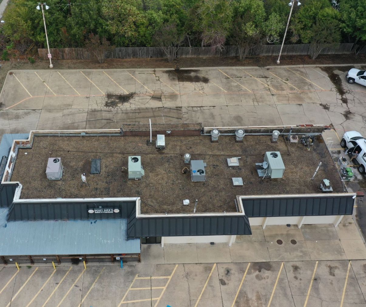 Overhead view of a commercial building with a flat roof, surrounded by a parking lot and trees.